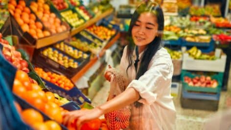 Young woman in a white shirt with a shopper bag choosing tangerines in a fresh vegetable and fruit store.