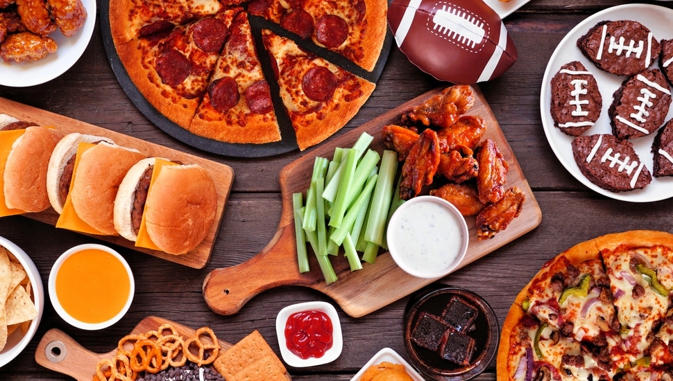 Super Bowl or football theme food table scene. Pizza, hamburgers, wings, snacks and sides. Overhead view on a dark wood background.