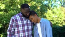 Teenager leans on his father's head, and his father consoles him.