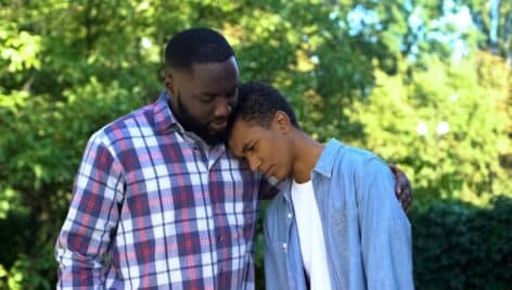 Teenager leans on his father's head, and his father consoles him.