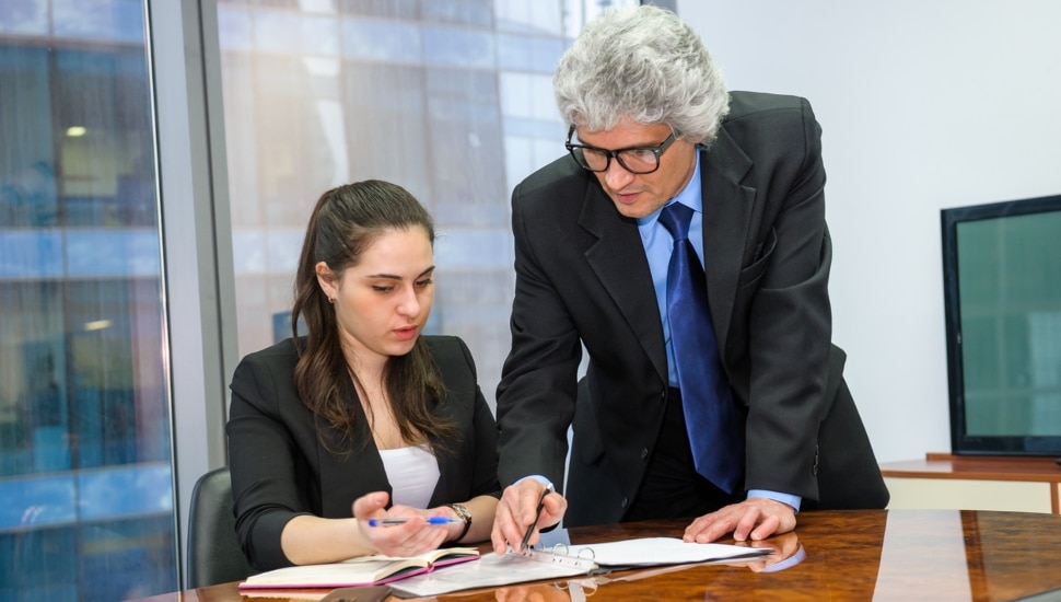 A woman sits at a desk with paperwork as a man guides her.
