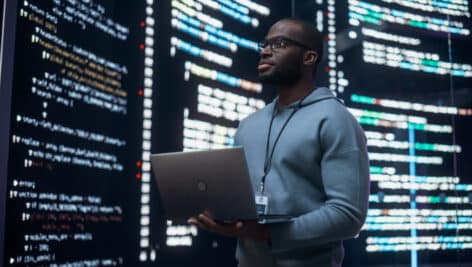 Man holding a laptop and looking at a big digital screen displaying back-end code lines.