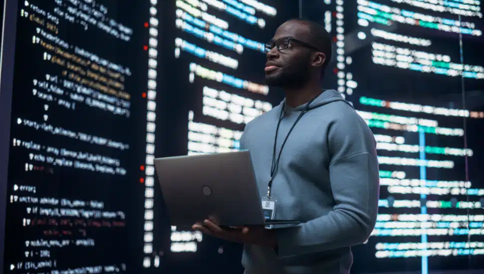 Man holding a laptop and looking at a big digital screen displaying back-end code lines.