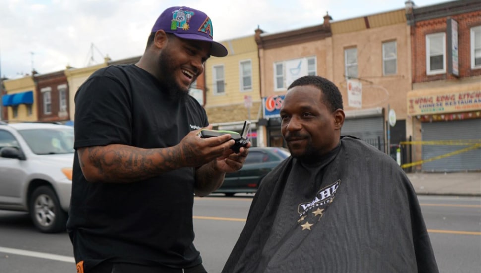 Josh Santiago smiles while giving man a haircut.