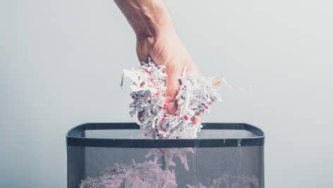 A hand is putting a bunch of shredded paper in a waste paper basket