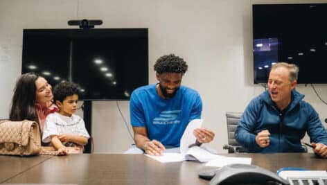 Joel Embiid smiles while signing contraction extension, with wife and son to his right and Josh Harris on his left.