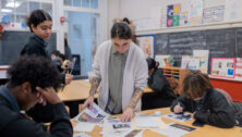 Students sit at their desks with teacher helping them out.