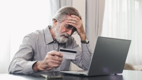 Upset senior elderly man holding credit card by laptop having trouble worry finance safety data or online payment security. Bank client concerned about problem with credit card, financial fraud threat