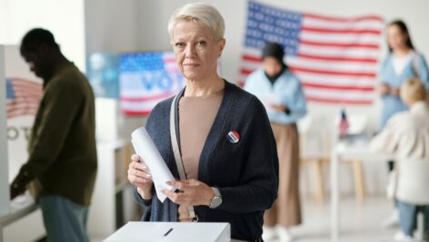 Mature blond female citizen of USA looking at camera while standing by box and holding ballot paper with her choice of candidate.