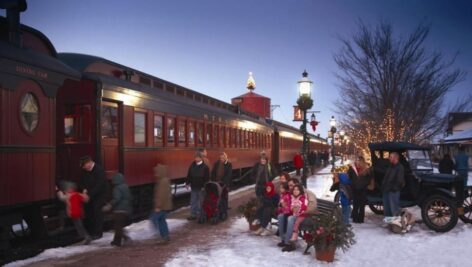 Families and children enjoy a festive evening at a holiday-themed train station, featuring a vintage train decorated with lights, snow-covered ground, classic cars, and seasonal decorations under a twilight sky.