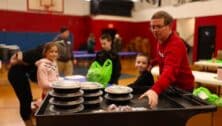 Man serving meals to those in need with children in the background.