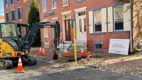 A small John Deere excavator at a construction site on a city street, surrounded by orange traffic cones and dug-up pavement. A sign nearby reads "Building a Better America" and includes logos for Aqua, the EPA, and the Bipartisan Infrastructure Law, indicating infrastructure work. The site is in front of a red brick building with white shutters and doors.