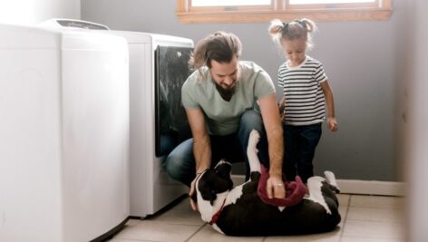 Father and daughter play with their dog while doing laundry.