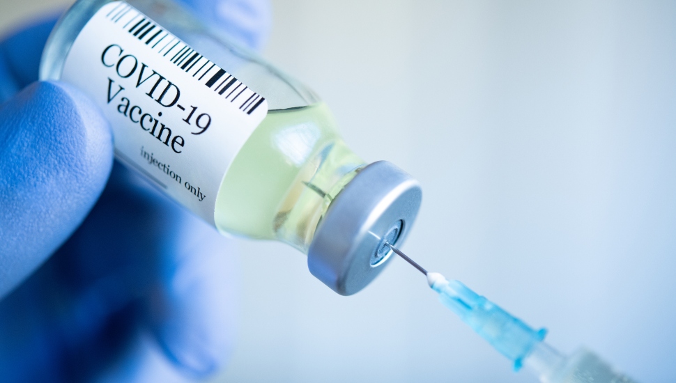 Close up of hand wearng protective glove and drawing up COVID-19 vaccine from glass phial bottle and filling syringe.