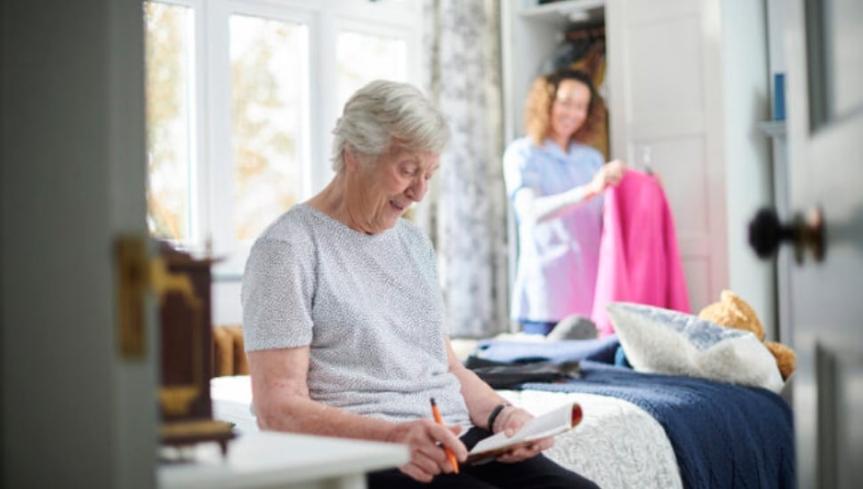 An elderly woman looks at a document while a helper hangs up clothes.