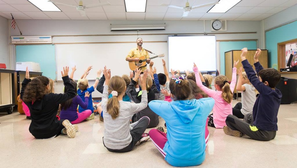 Kids sitting with arms raised while instructor plays a guitar.