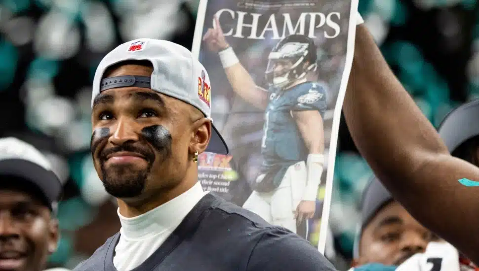 Jalen Hurts smiling with Eagles Super Bowl hat and shirt on. A newspaper with "Champs" written on it in the background.