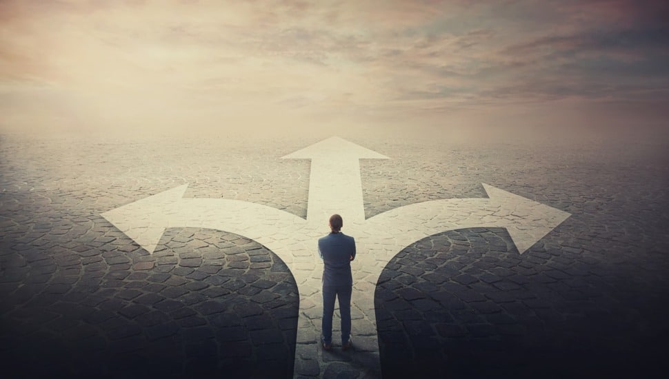 Doubtful businessman stands in front of a crossroad with road split in three different ways as arrows show opposed courses.