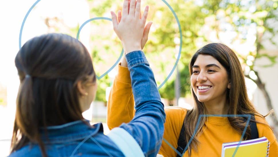 Two female college students holding books, smiling, and high-fiving.