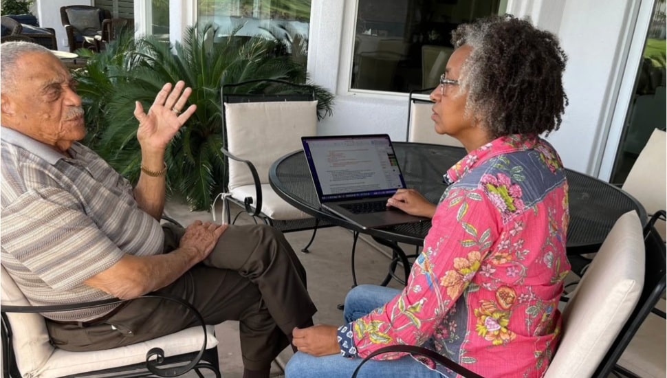 George E. Johnson (left) sits and talks with Hilary Beard (right) with a laptop in between them on the table.
