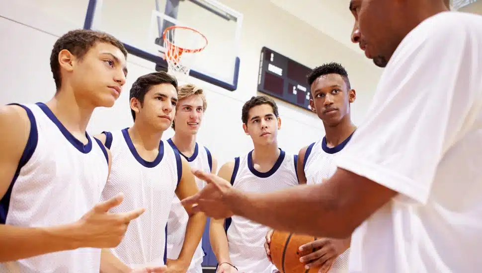 A group of four high school basketball players being addressed by their coach on the sidelines.