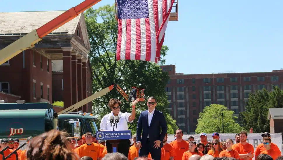 Gov. Shapiro and another official speaking at a podium in front of workers, with a crane and American flag in the background.