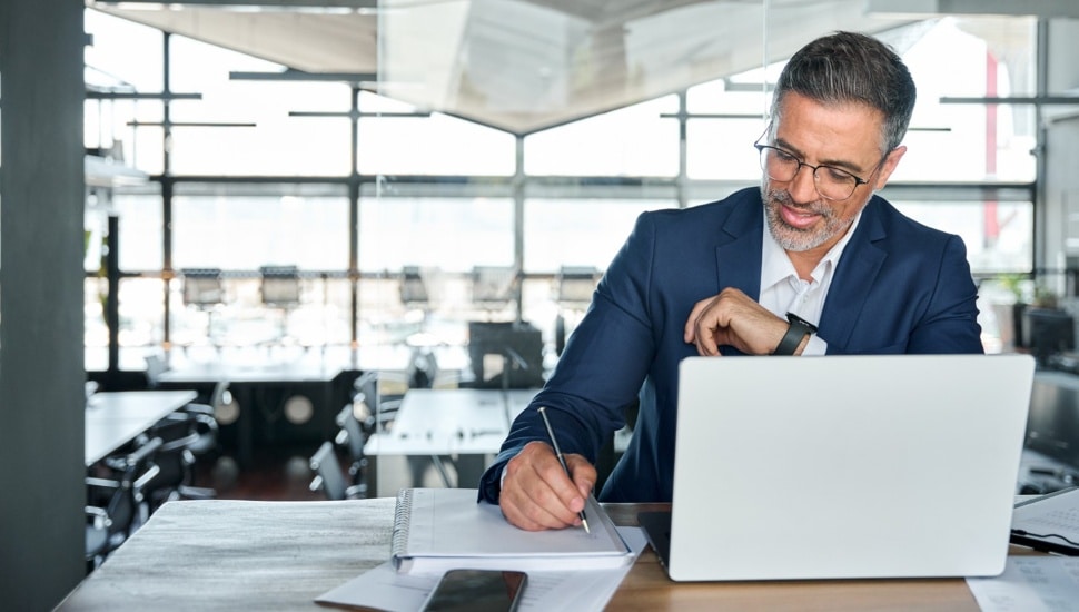 Middle-aged businessman working on laptop computer in office writing notes.