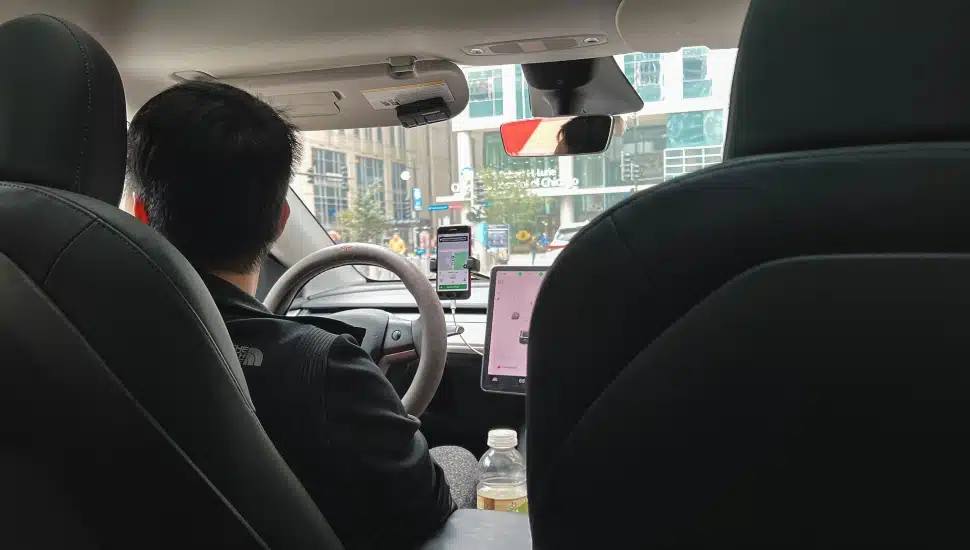 Passenger point of view sitting in the back seat of a ride share vehicle. A male driver sits in the front of the car and he holds the steering wheel.