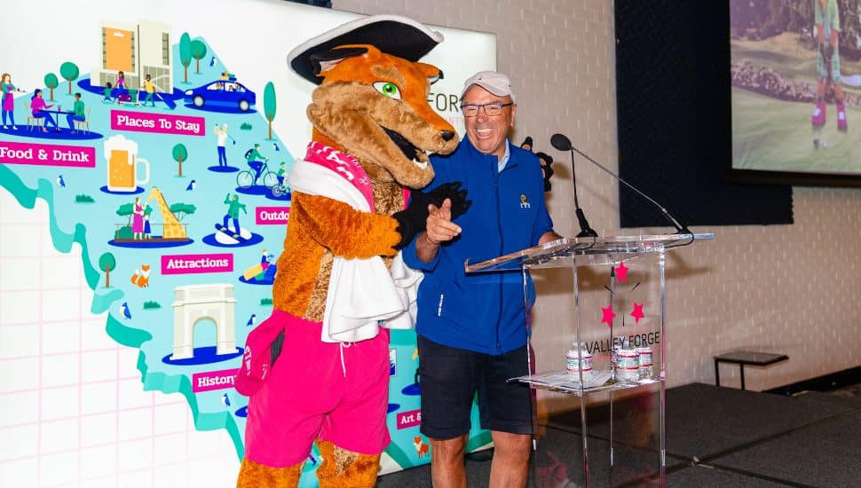 Mike Bowman wears casual golf attire and shares a laugh on stage with Monty the Fox, the Valley Forge Tourism mascot, during the 2025 Travel Rally event at Union League Liberty Hill, with a colorful tourism map in the background.