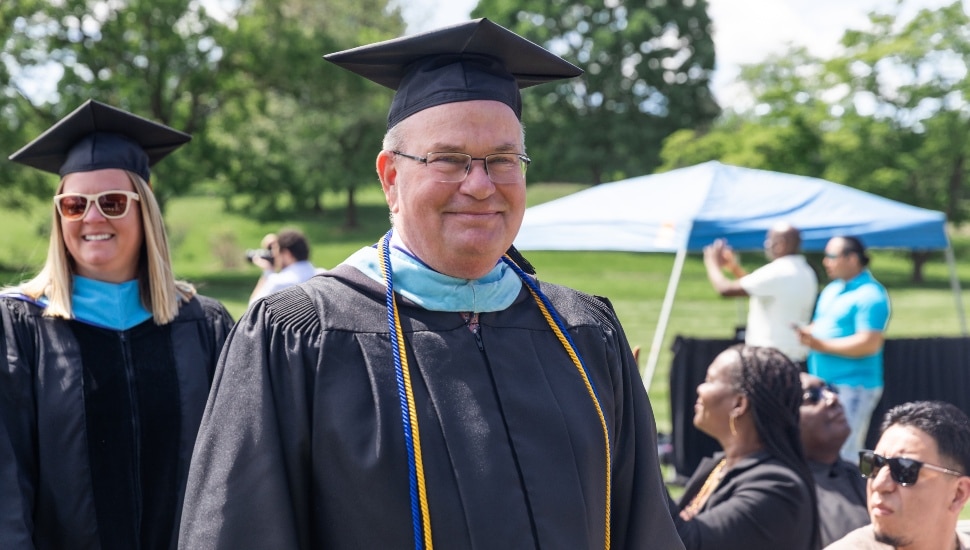 Older white male prep school administrator smiles in cap and gown.