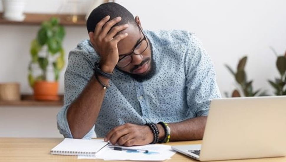 Young black male with beard and glasses sits frustrated at a laptop computer.