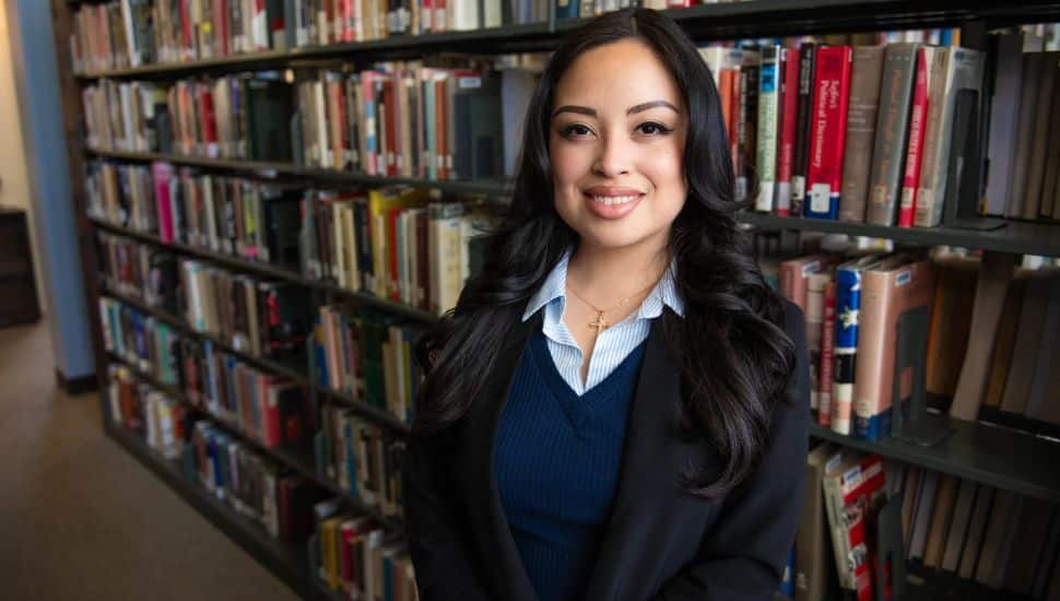 Headshot of Kimberly Alfaro Jimenez in a library setting, with a background of rows of books. She has long dark hair and wears navy and black, smiling.