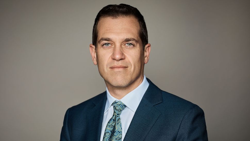 Portrait of Michael Vennera in a blue suit and patterned tie, looking directly at the camera with a neutral expression against a plain background.