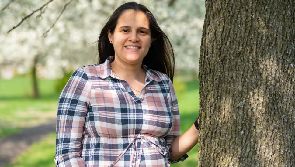 Rocio Rodriguez: Smiling woman with long dark hair wearing a pink, navy, and white plaid shirt standing outdoors next to a tree trunk, with a blurred background of green grass and blooming white flowers.