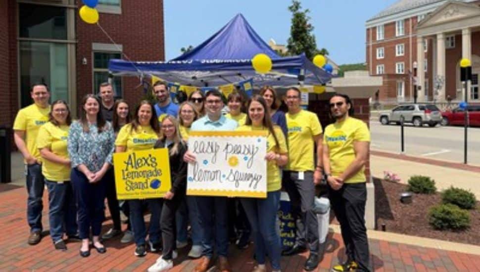 A group of S&T Bank employees wearing yellow shirts pose in front of a lemonade stand tent with signs supporting Alex’s Lemonade Stand Foundation during a community fundraising event.