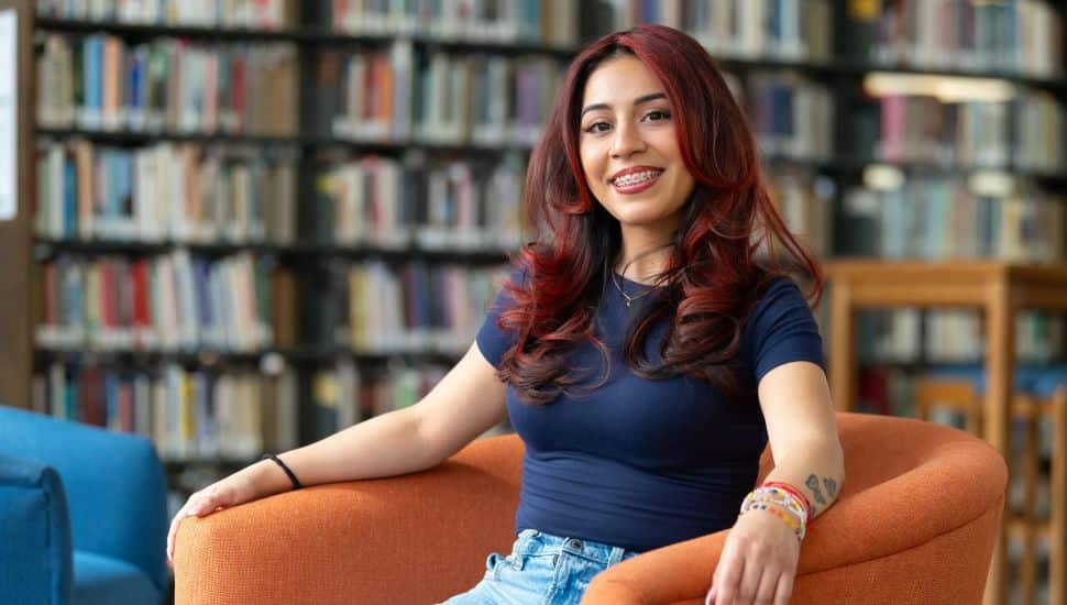 A young woman with long, wavy dark red hair and braces is sitting comfortably in an orange armchair in a library. She is smiling and wearing a navy blue fitted t-shirt and light blue jeans. She has colorful bracelets on one wrist and a small tattoo on her forearm. Bookshelves filled with books are visible in the background, giving the setting a cozy, academic atmosphere.