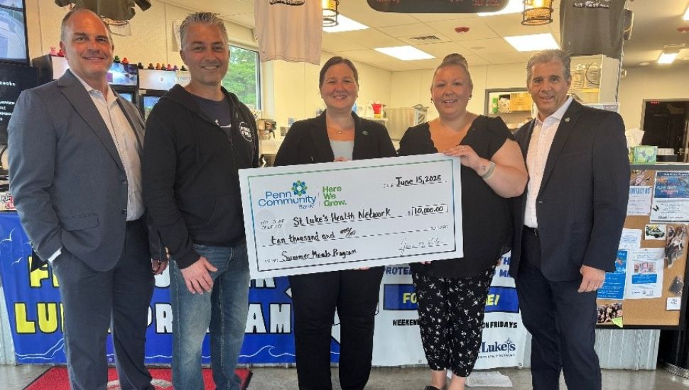 Left to Right: Dennis Pfleiger, President of St. Lukes University Health Network – Quakertown Campus; Pastor Dave Kratz, Executive Director – Free Fall Action Park; Kelly Boyle – Manager of Corporate Social Responsibility & Government Affairs, Penn Community Bank; Jessica Althouse – Financial Center Manager, Penn Community Bank; Paul Gregg – Regional Retail Manager, Penn Community Bank. The check is from Penn Community Bank and made out to St. Luke’s Health Network for $10,000.00, dated Jun. 13, 2025, and designated for the Summer Meals Program. The setting appears to be a casual community space or café, with promotional posters and merchandise in the background.