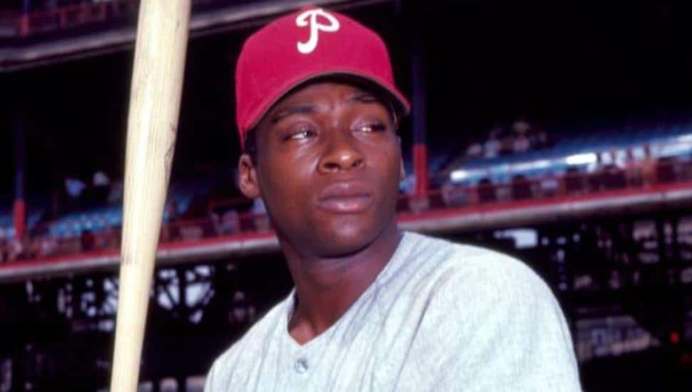 Dick Allen is holding a bat on the field in a red Phillies hat.