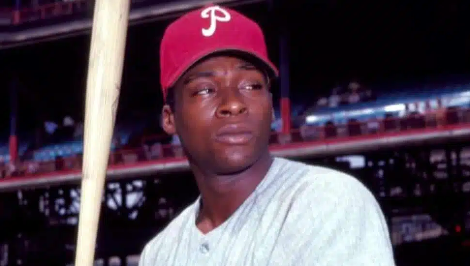 Dick Allen is holding a bat on the field in a red Phillies hat.