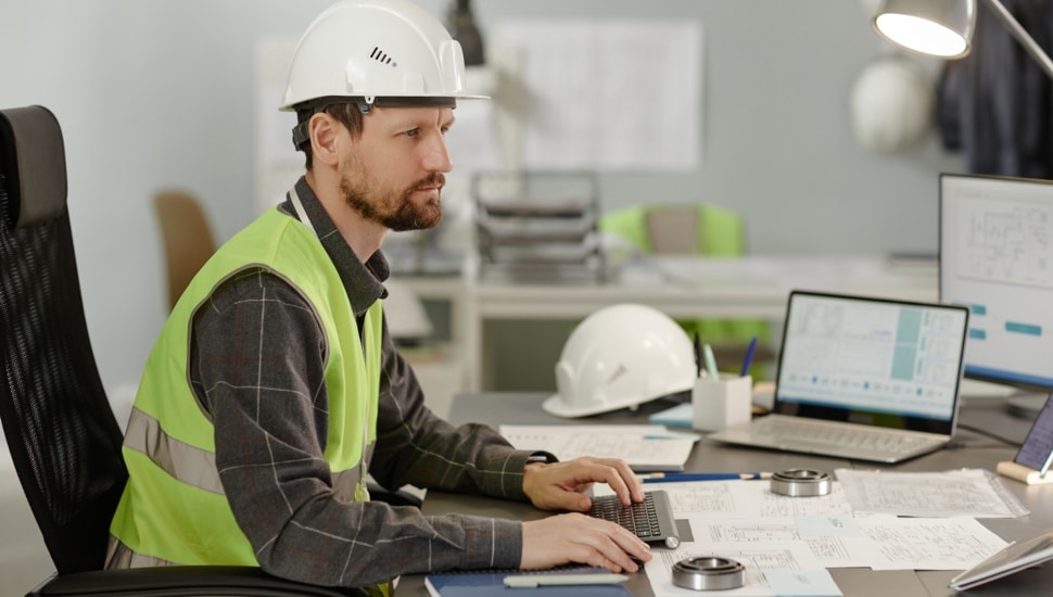 Side view portrait of bearded construction engineer wearing hardhat at workplace in office and using computer