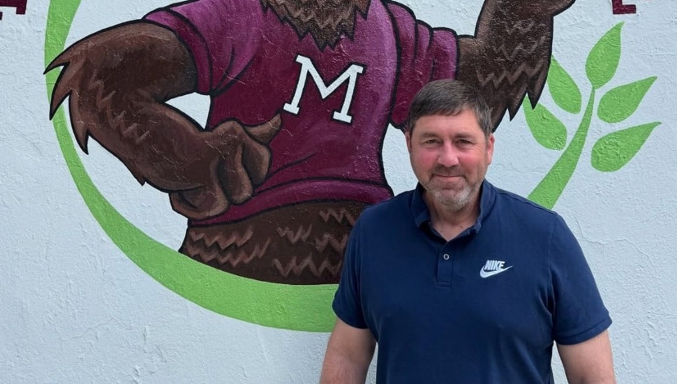 Middle-aged white male in blue polo shirt poses for picture in front of painting of school mascot.