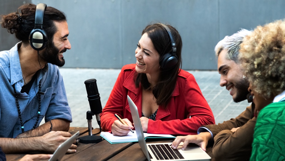 Smiling young influencers record podcast outdoors using microphone, headphones and laptop.