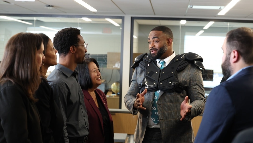 Football player wearing suit and shoulder pads talks to bank employees.
