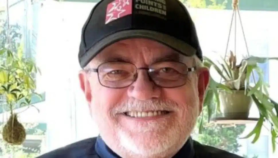 Smiling older man with glasses, a gray beard, and a black "Turning Points for Children" cap, standing indoors with hanging plants in the background.