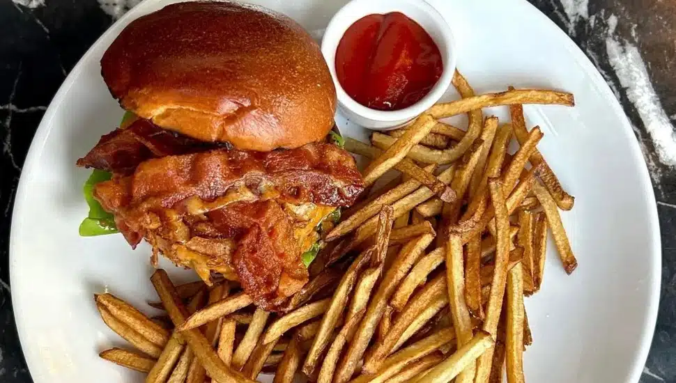 A burger topped with bacon, cheese, and lettuce, surrounded by fries and a cup of ketchup.