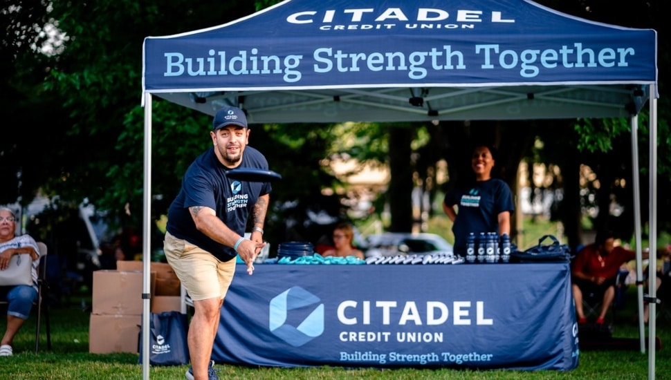 Credit union employee throws frisbee outdoors.