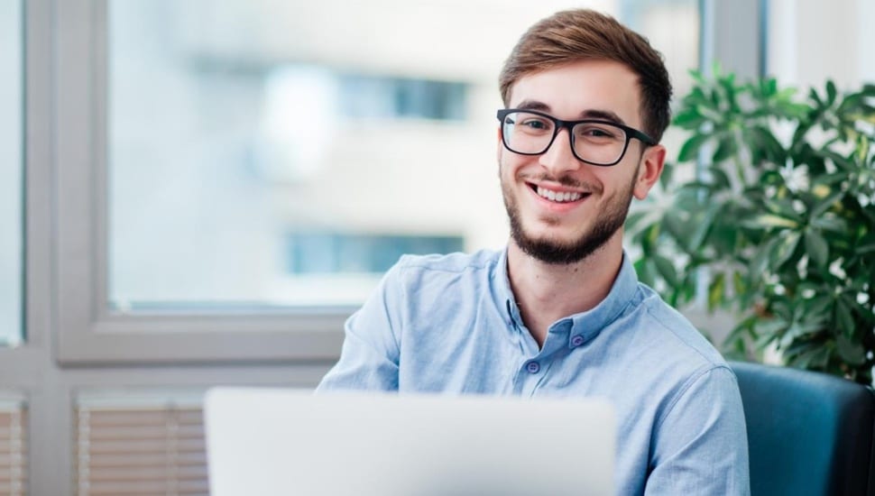 Male college student with glasses smiles while seated at desk.