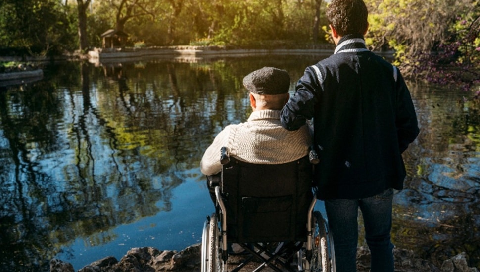 Son stands with his wheelchair-bound father at the edge of a pond.