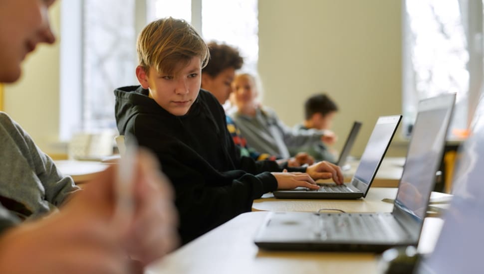 A boy sits in a group working at laptops, staring off into space.
