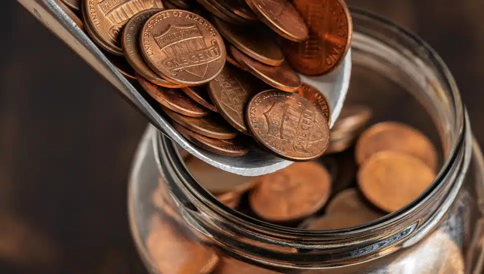 A metal scoop pouring U.S. pennies into a glass jar.